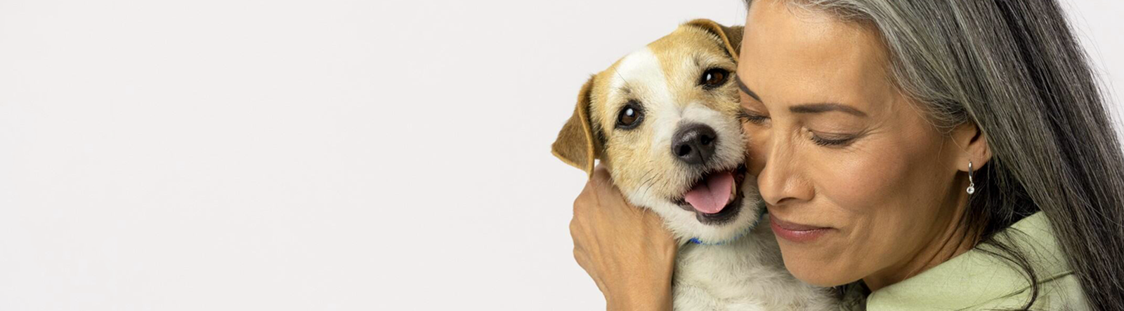 A woman with long grey hair smiling and hugging a small brown and white dog. The dog has its mouth open in a happy expression against a plain white background.

