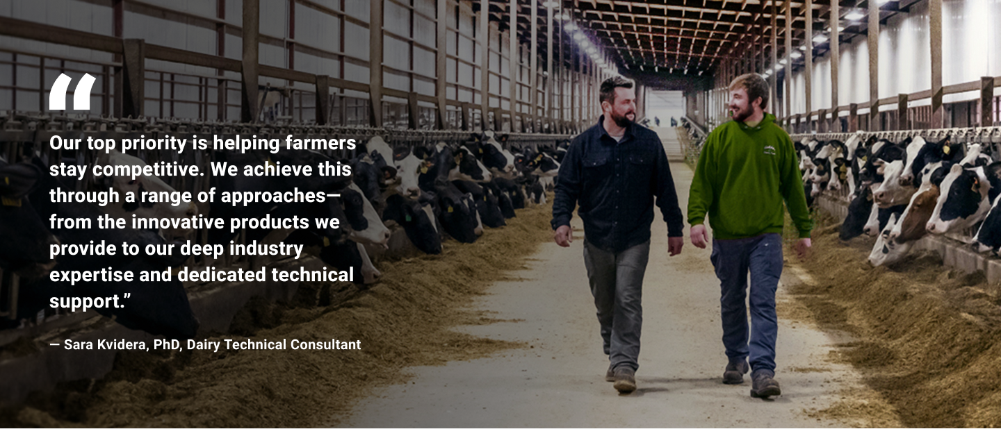 Two men walk through a modern dairy barn with rows of Holstein cows eating from feed troughs, alongside a quote from Sara Kvidera, PhD, Dairy Technical Consultant, about helping farmers stay competitive.