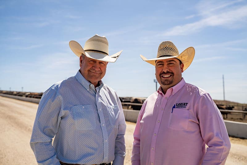 Portrait of two smiling cattle producers wearing cowboy hats and button-down shirts, standing in front of a livestock feedlot under a clear blue sky.