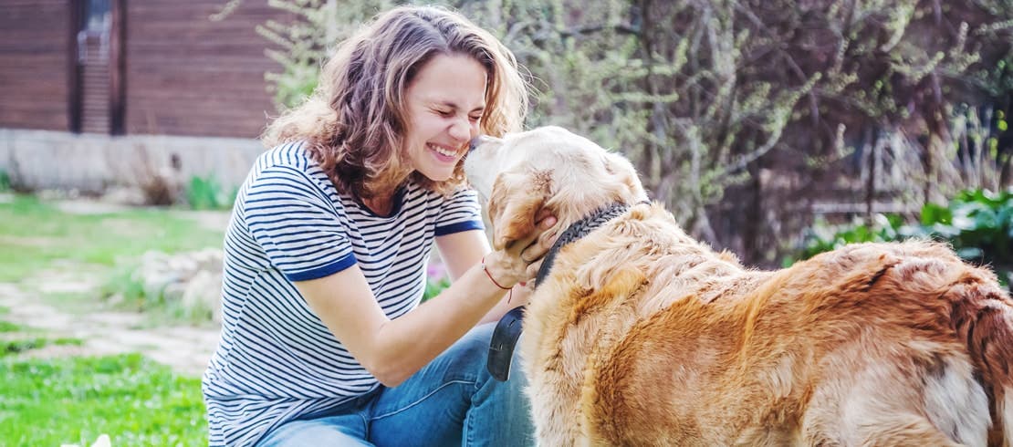 A very happy woman bent down petting the face of her senior golden retriever in the backyard.